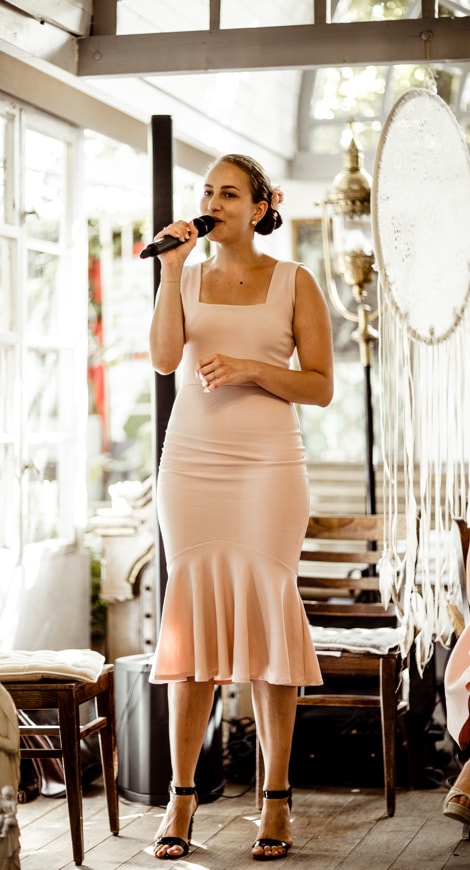 Wild Hearts, Sam, Traurednerin, Sängerin, auf einer Hochzeit in einem rosa Kleid. Im Hintergrund sind einige alte Holzstühle und ein weißes Fenster zu sehen, großer Traumfänger, Macramee, Rosa